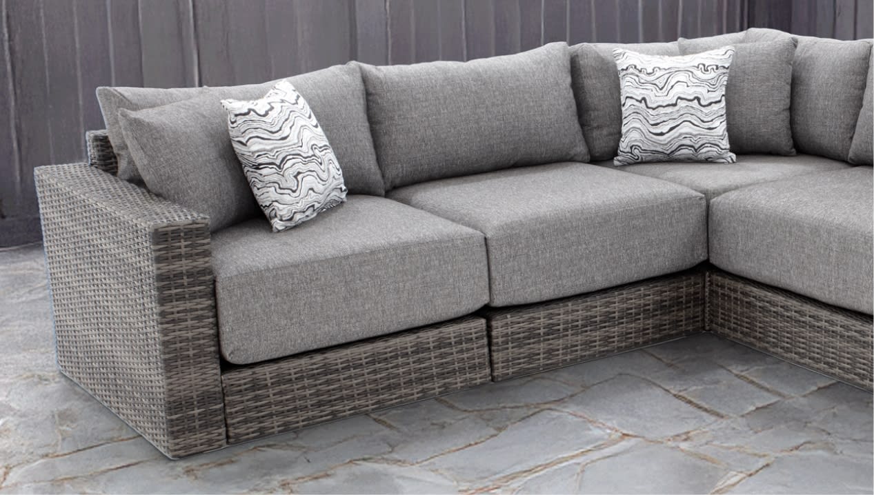 Modern L-shaped outdoor sectional sofa with gray woven rattan base, light gray fabric cushions, and two white and black patterned throw pillows on stone tile flooring.