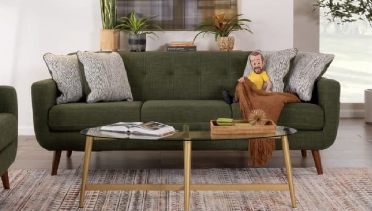 A mid-century modern living room features a green sofa with patterned cushions, Little Bob , and a warm throw. A glass coffee table holds a book and decor.