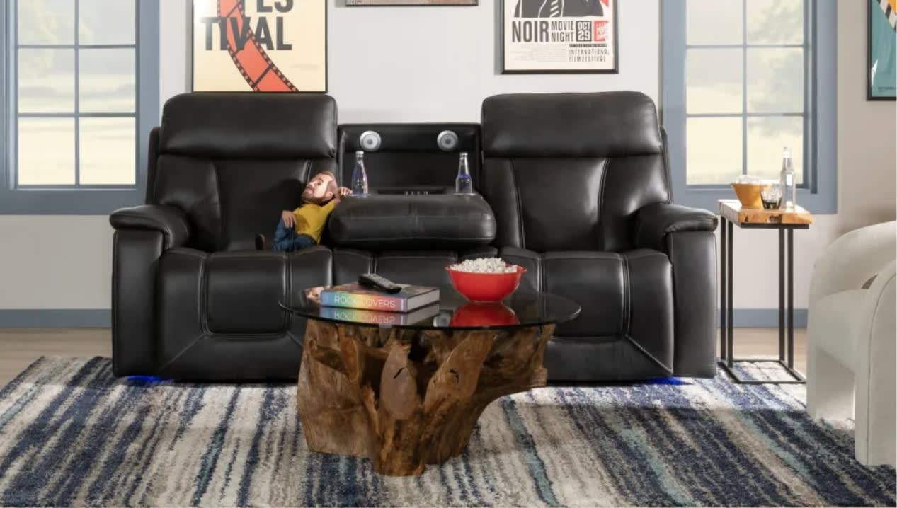 Black leather reclining loveseat with center console featuring cup holders and storage, paired with a rustic wooden coffee table with glass top on a striped area rug.