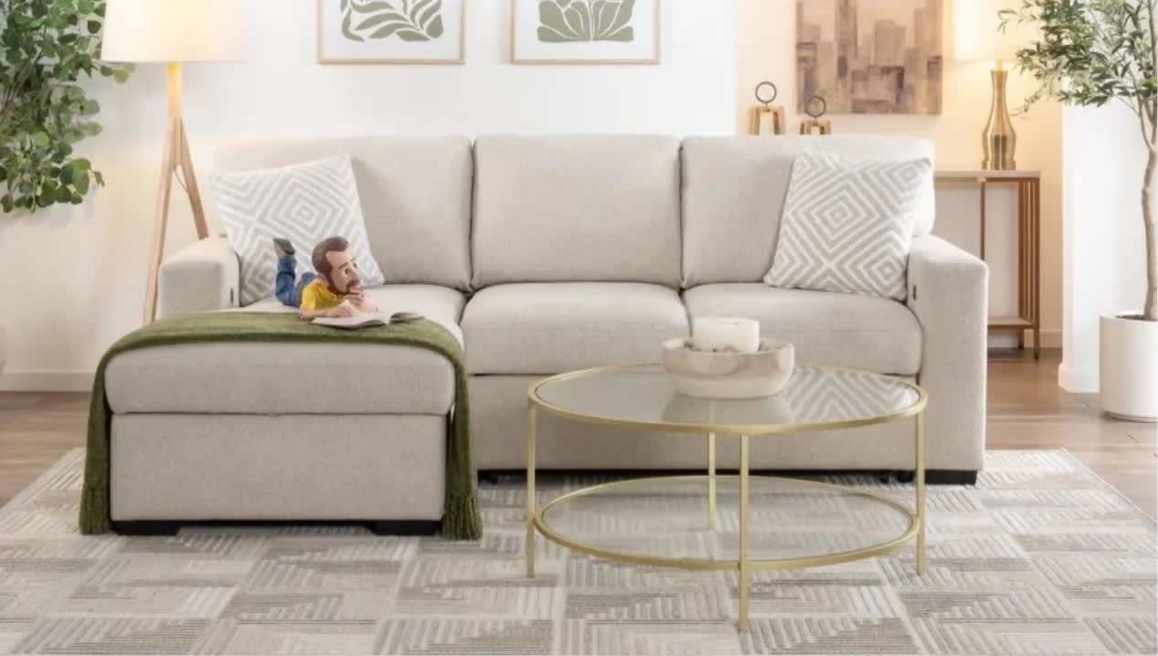 A cozy living room features a light beige sofa with geometric pillows. Little Bob reads on the sofa under a green throw. A round glass coffee table sits on a patterned rug.