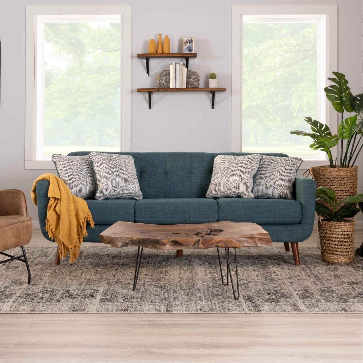 Modern living room setup with a teal fabric sofa featuring wooden legs, patterned gray cushions, a natural wood coffee table with metal hairpin legs, and woven plant baskets.