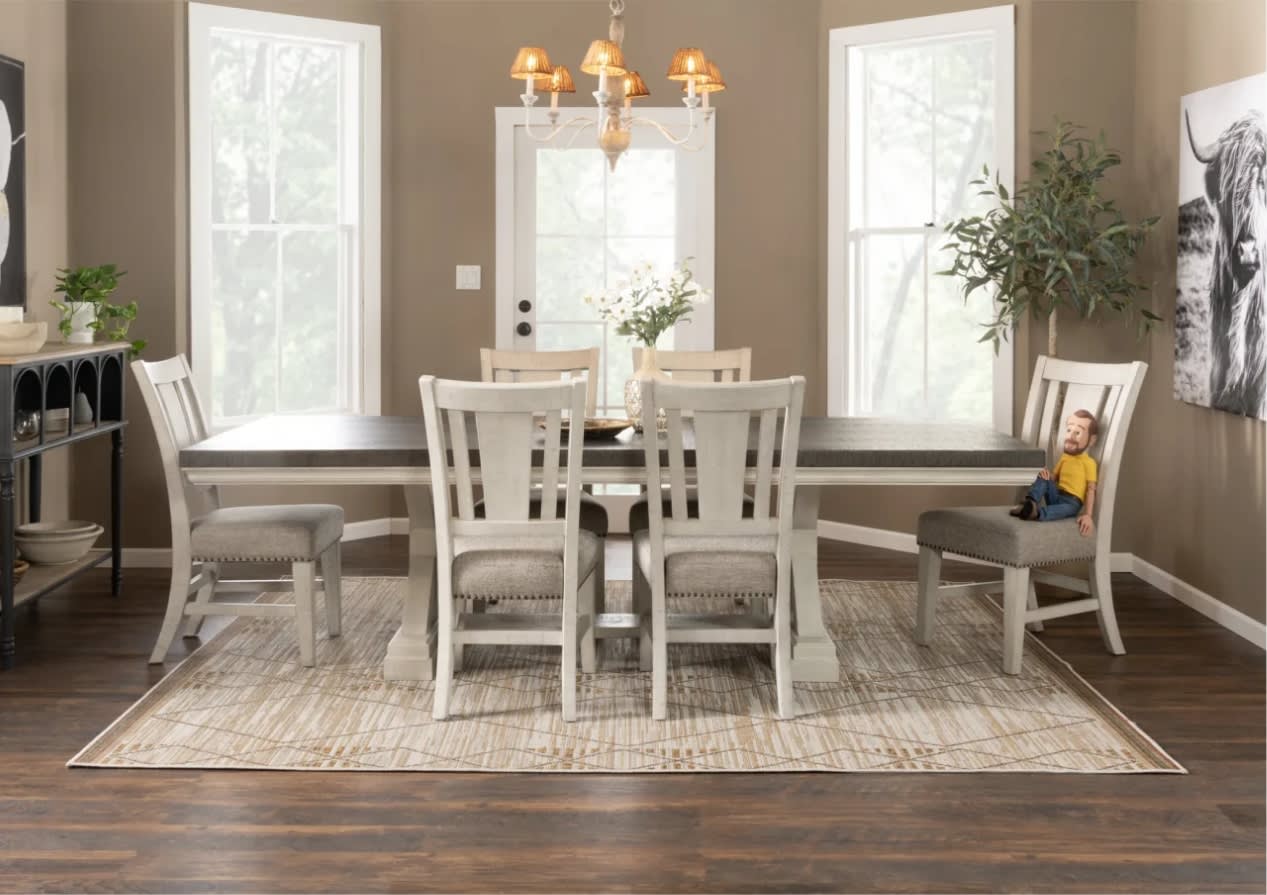 Dining room with a long table, six chairs on a beige rug, and large windows. A chandelier hangs above; a plant and doll sit on chairs, offering a cozy ambiance.