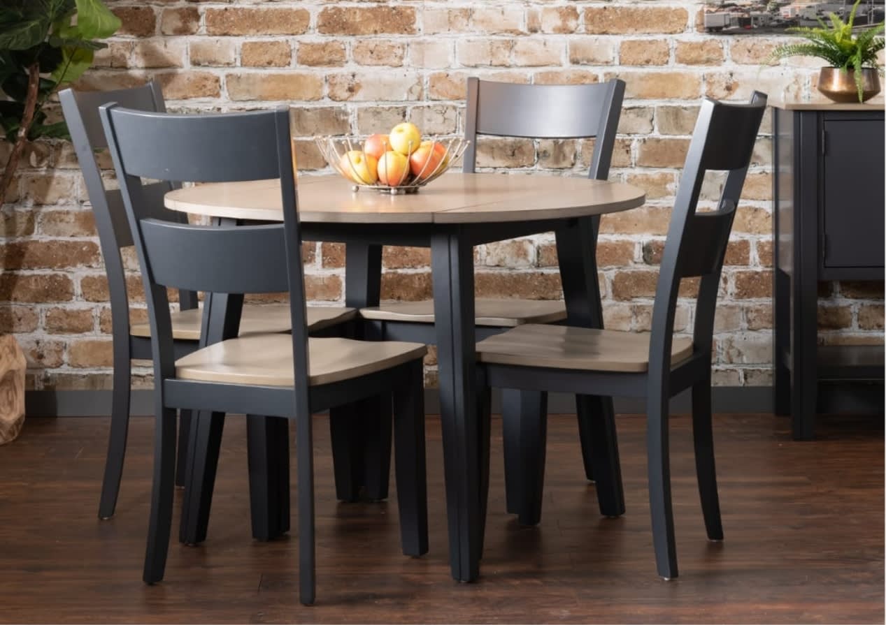 A round wooden dining table with four black chairs, set against a rustic brick wall. A fruit bowl with oranges sits at the center, creating a cozy ambiance.