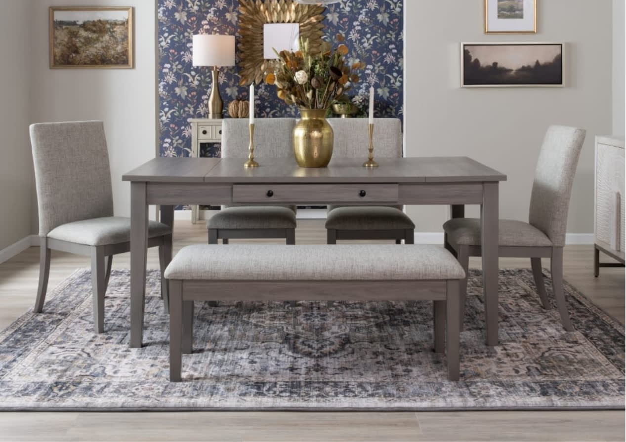 Elegant dining room with a gray table, cushioned chairs, and a matching bench on a patterned rug. A gold vase, floral wallpaper, and framed art enhance the decor.