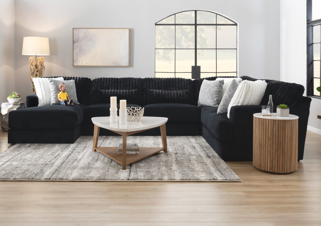 Modern living room with a large black sectional sofa featuring textured and white throw pillows, a wooden coffee table with marble top, and a wooden side table with a small plant and bottle.