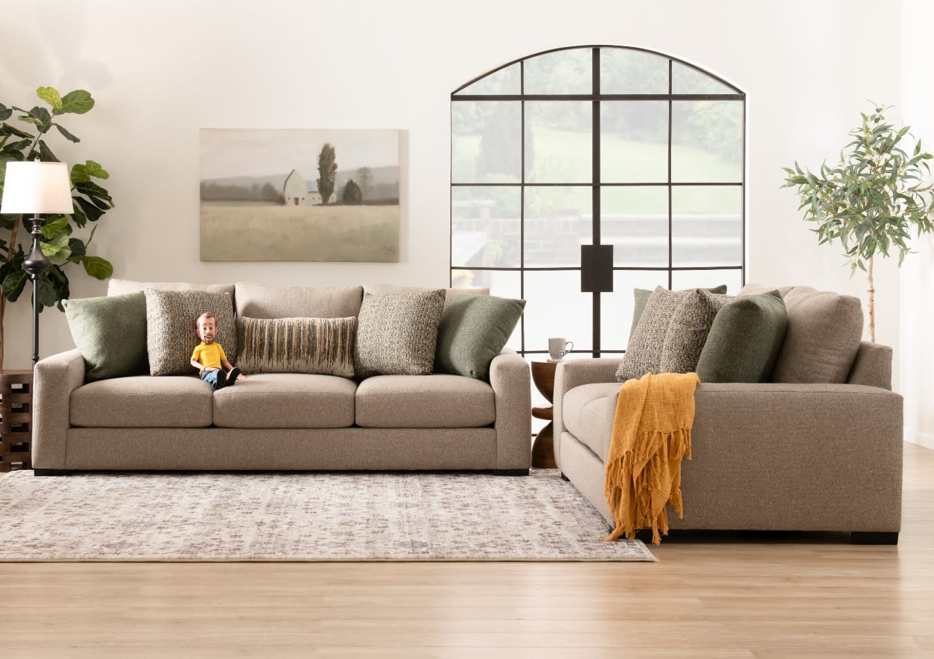 Modern living room with two beige fabric sofas featuring multiple textured cushions in green, grey, and brown tones, a mustard throw, a wooden side table with a cup, a floor lamp, and a large arched window with black grid framing.