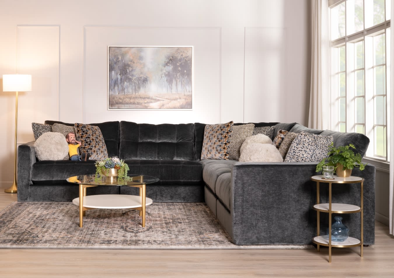 Dark gray velvet L-shaped sectional sofa with multiple patterned and textured throw pillows, gold and glass round coffee table, side table with plant, floor lamp, and large window.