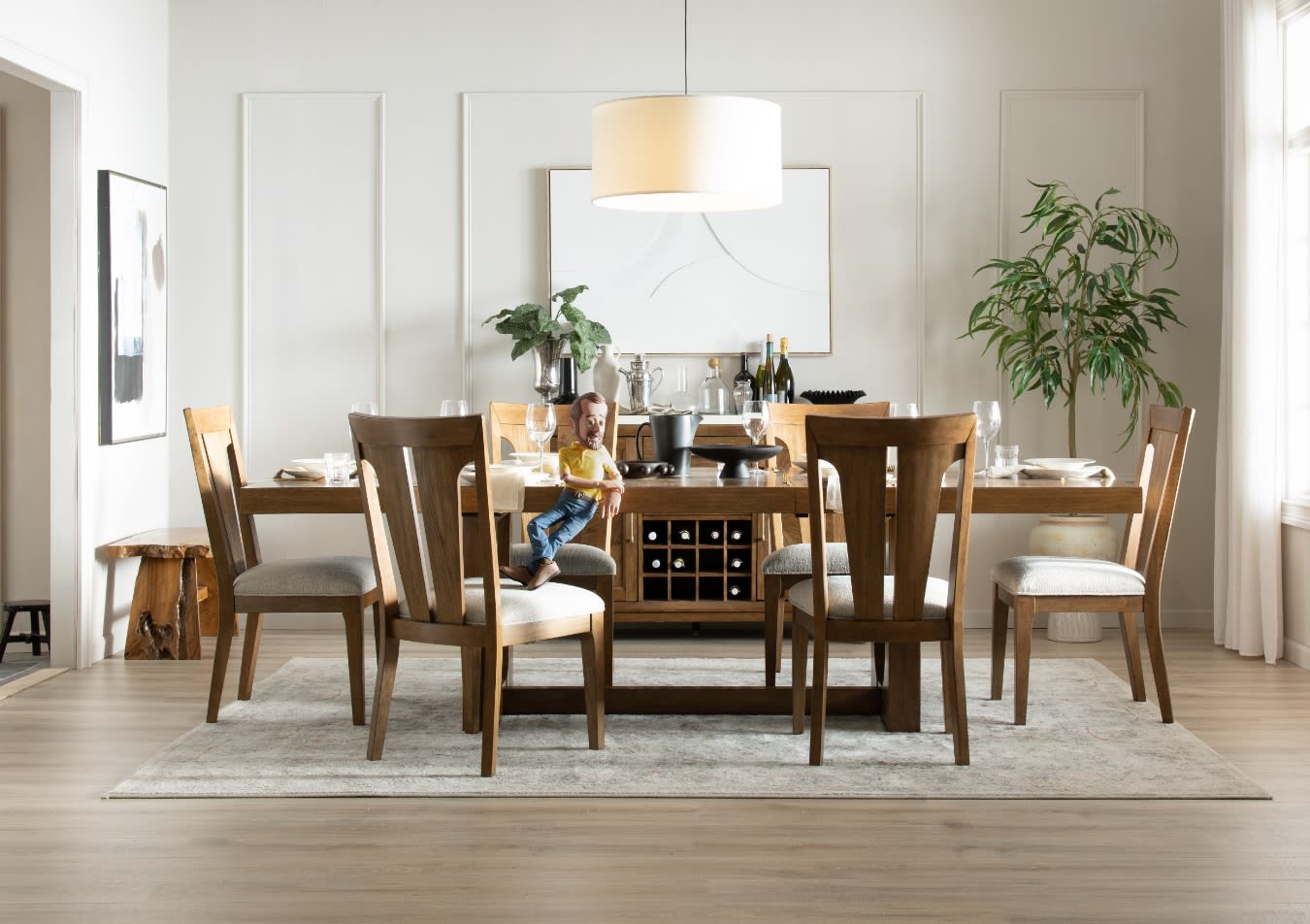 Modern dining room with a wooden table and six cushioned chairs on a light gray rug, featuring a wine rack, pendant light, and potted plant.
