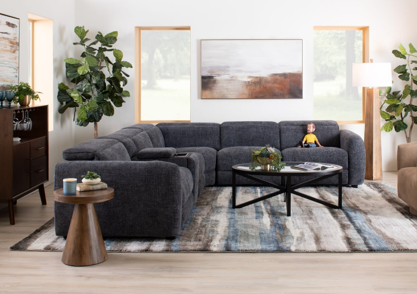 Dark gray fabric sectional sofa with plush cushions and adjustable headrests in a modern living room featuring a round glass coffee table, wooden side table, abstract wall art, and large leafy plants.