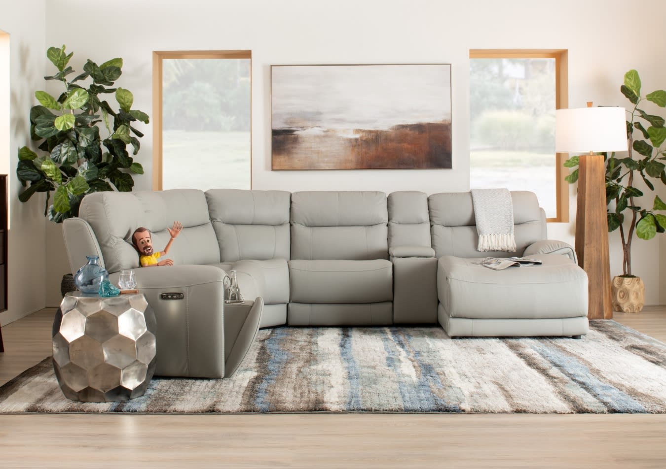 Modern living room setup with a light gray leather sectional sofa featuring recliners, a textured area rug, geometric metal side table, and wooden floor lamp.