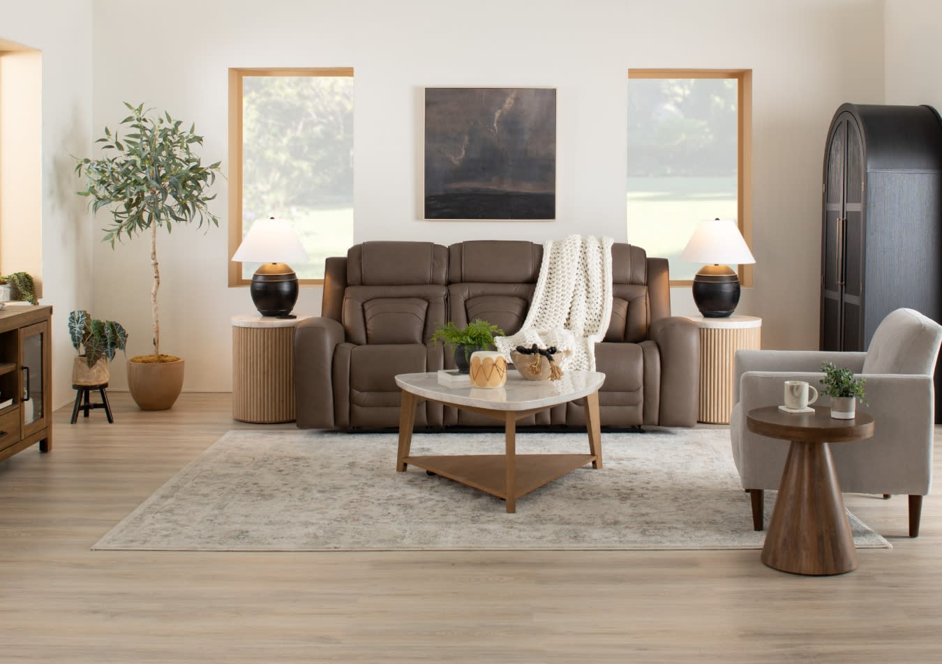 Modern living room with a brown leather sofa, beige armchair, wooden coffee table with marble top, side tables with black lamps, and decorative plants.