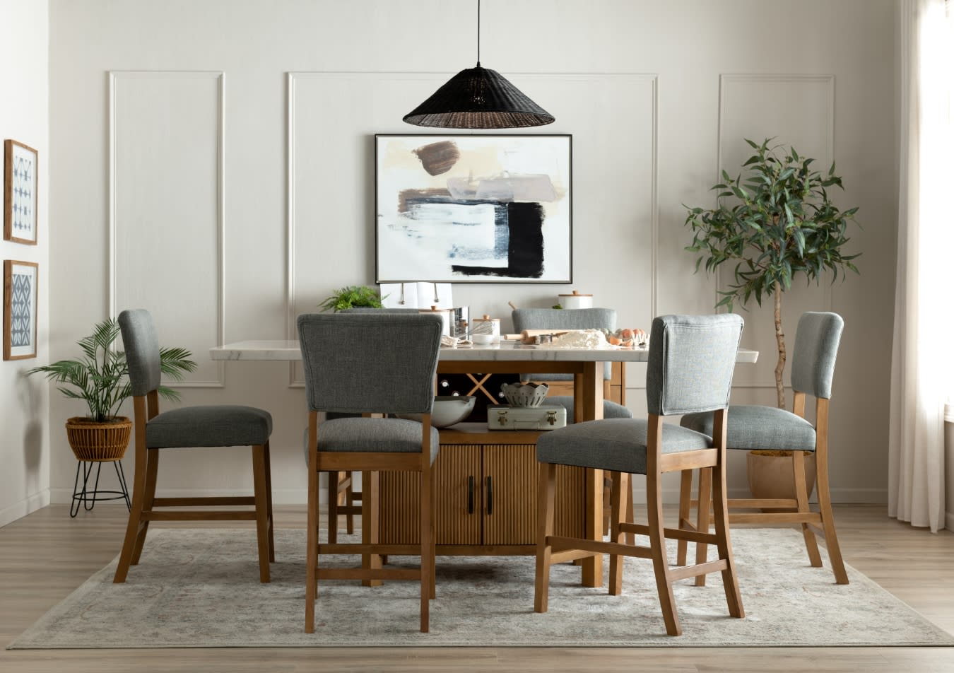 Dining room setup with a rectangular wooden table and six gray upholstered chairs on a patterned rug, featuring modern abstract wall art and indoor plants.