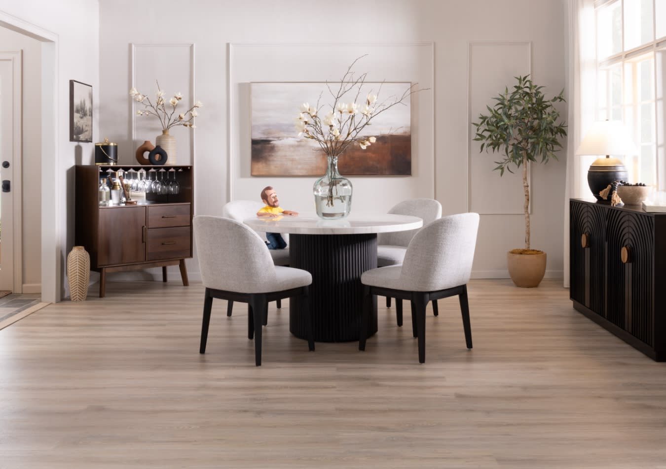 Modern dining room with a round white marble table, four light gray upholstered chairs with black legs, a large glass vase with branches centerpiece, wooden sideboard with glassware, and a black cabinet with a lamp and potted tree, featuring light wood flooring and neutral wall paneling.