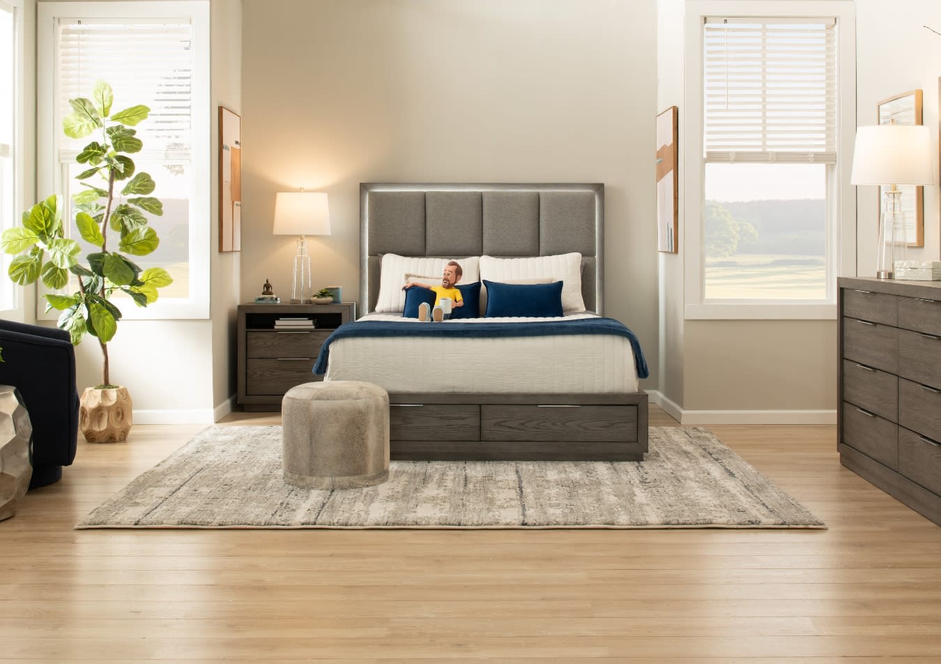 Modern bedroom with a gray upholstered bed, white and navy bedding, wooden nightstand and dresser, beige area rug, and natural light from windows.