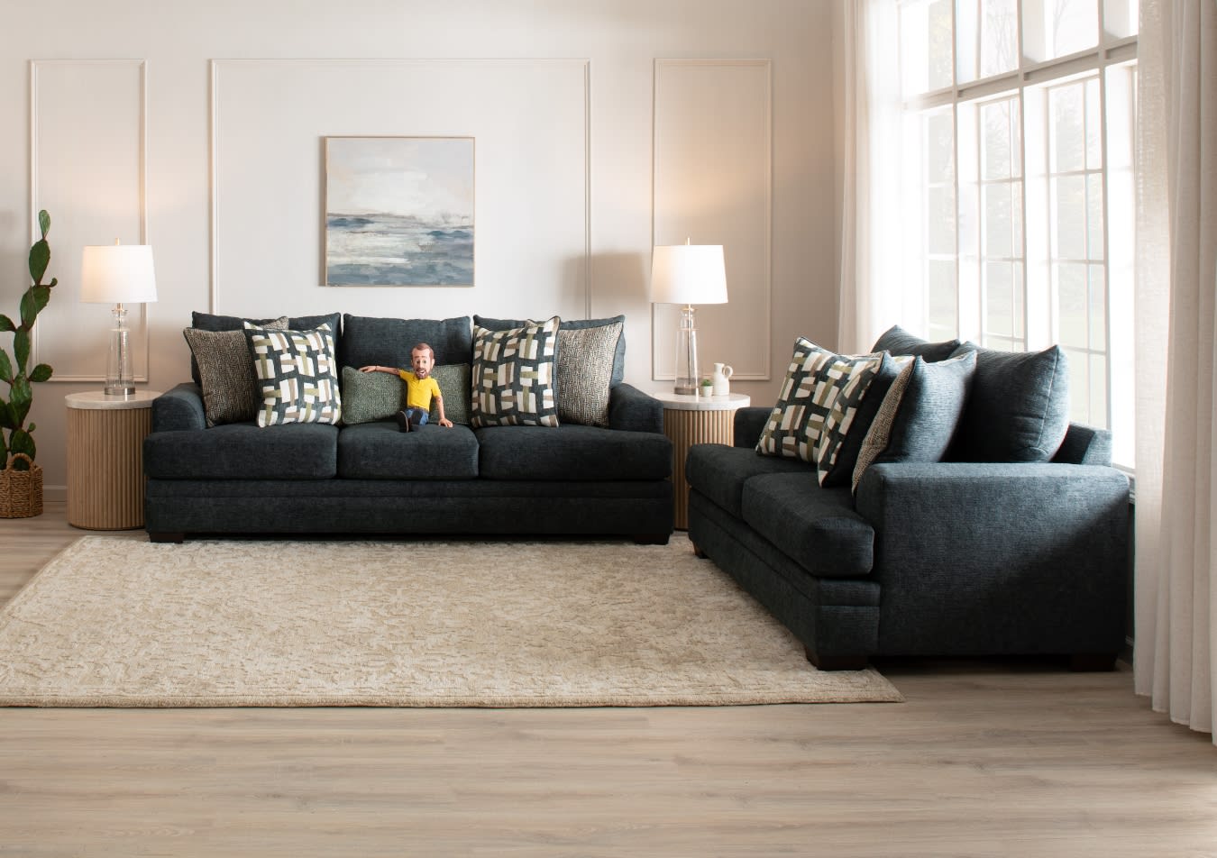 Modern living room with dark gray fabric sofas featuring patterned throw pillows, beige area rug, wooden side tables with glass lamps, and large windows.