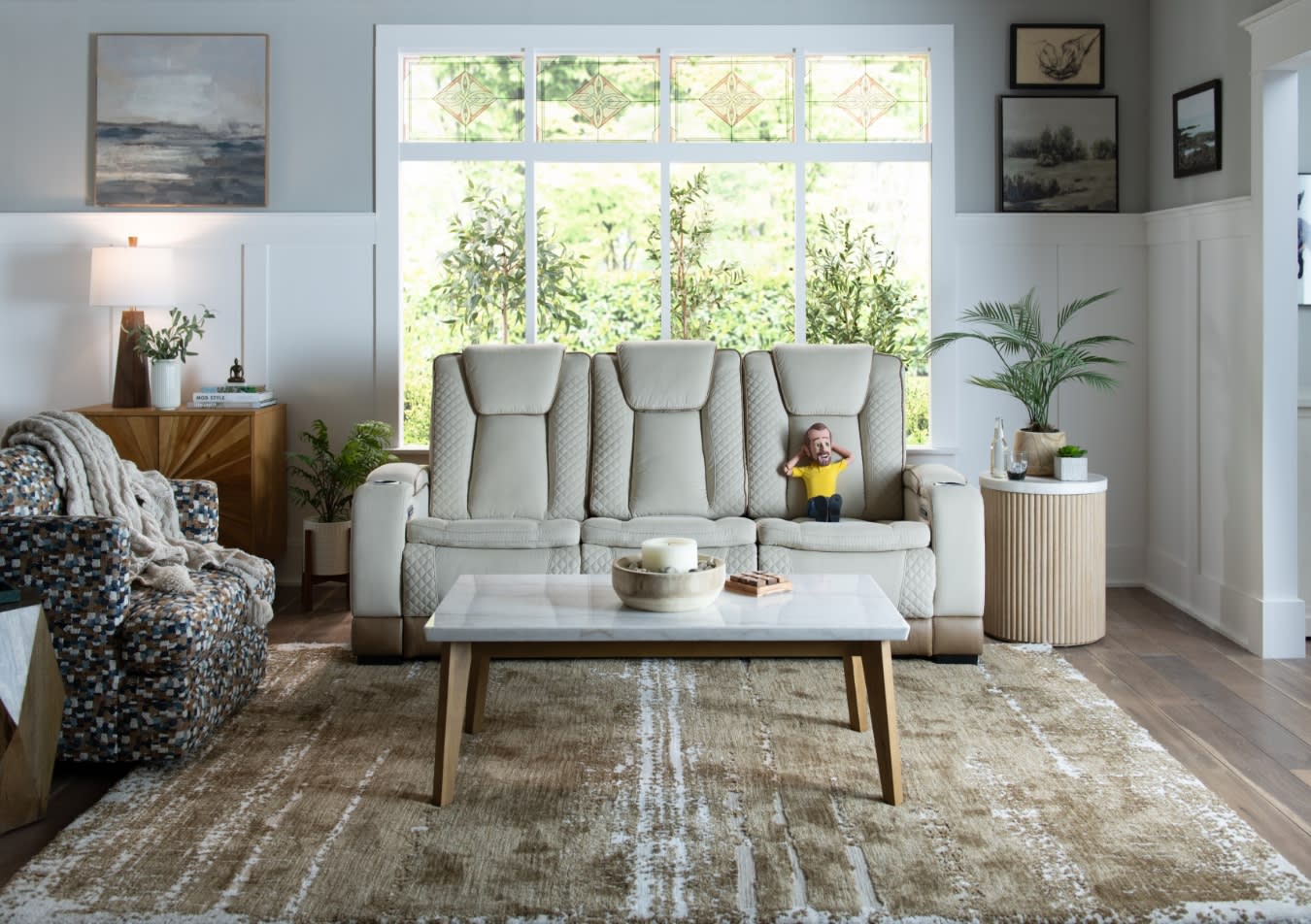 Modern living room with a beige quilted leather sofa, patterned armchair, marble coffee table, wooden flooring, and large window with stained glass accents.