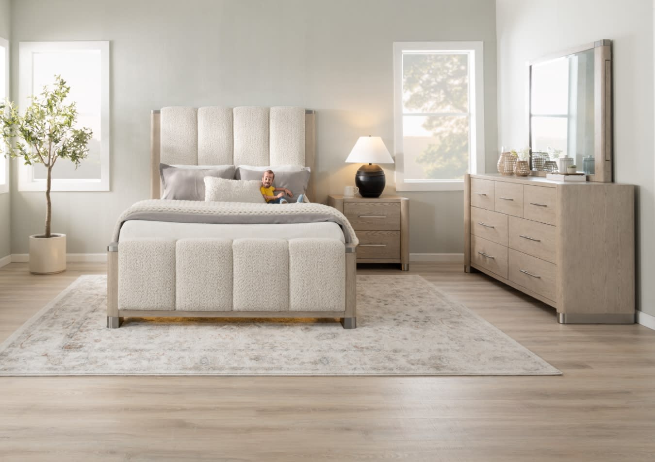 Modern bedroom with a beige upholstered bed featuring vertical paneling, matching nightstand with lamp, light wood dresser with mirror, and a neutral area rug on light hardwood floors.