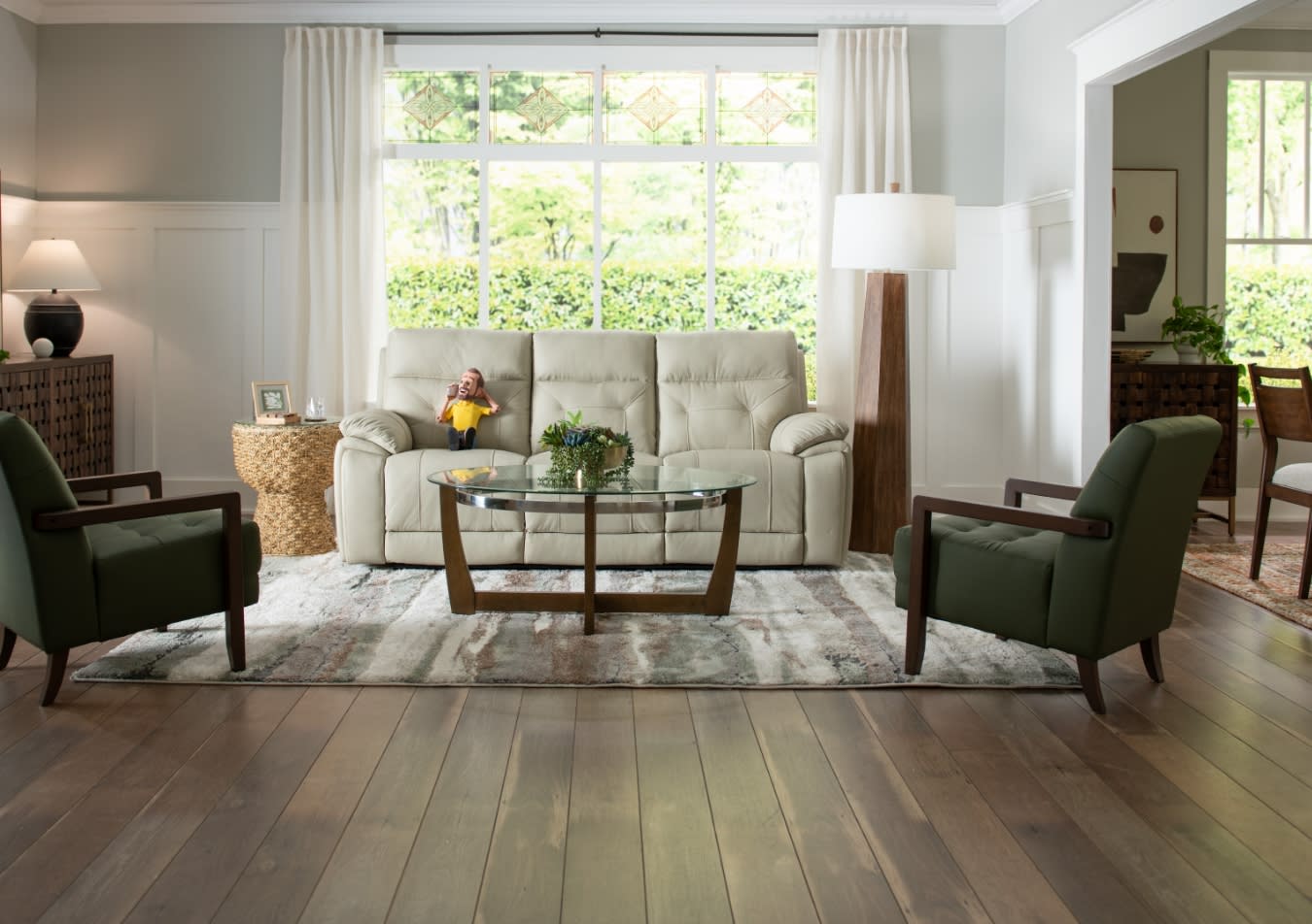 Modern living room with beige leather sofa, two green upholstered armchairs, glass coffee table, wooden side tables, and large window with white curtains.