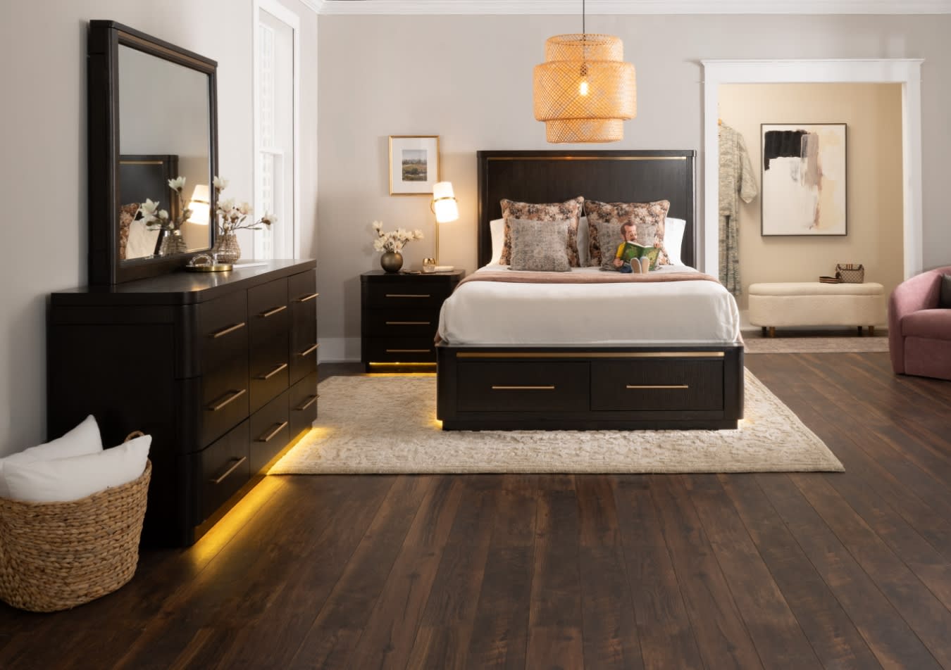 Modern bedroom with dark wood furniture including a bed with storage drawers, matching dresser and nightstand with brass handles, beige walls, hardwood floors, and warm ambient lighting.