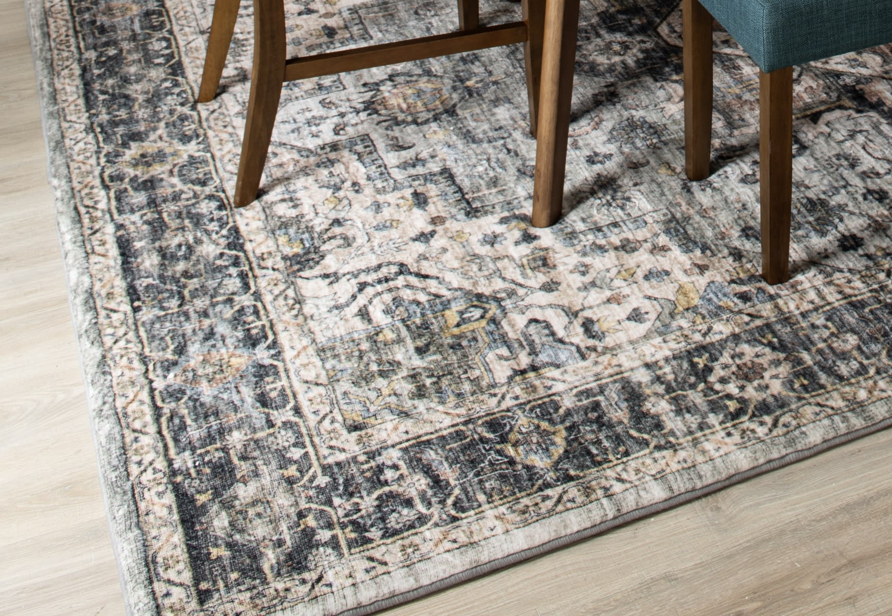 Vintage-style area rug with distressed floral patterns in muted gray, beige, and blue tones, placed under wooden dining table legs on light hardwood floor.