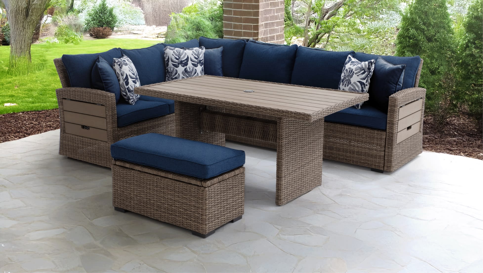 Outdoor patio furniture set with brown wicker frame, navy blue cushions, floral accent pillows, rectangular table, and matching cushioned ottoman.