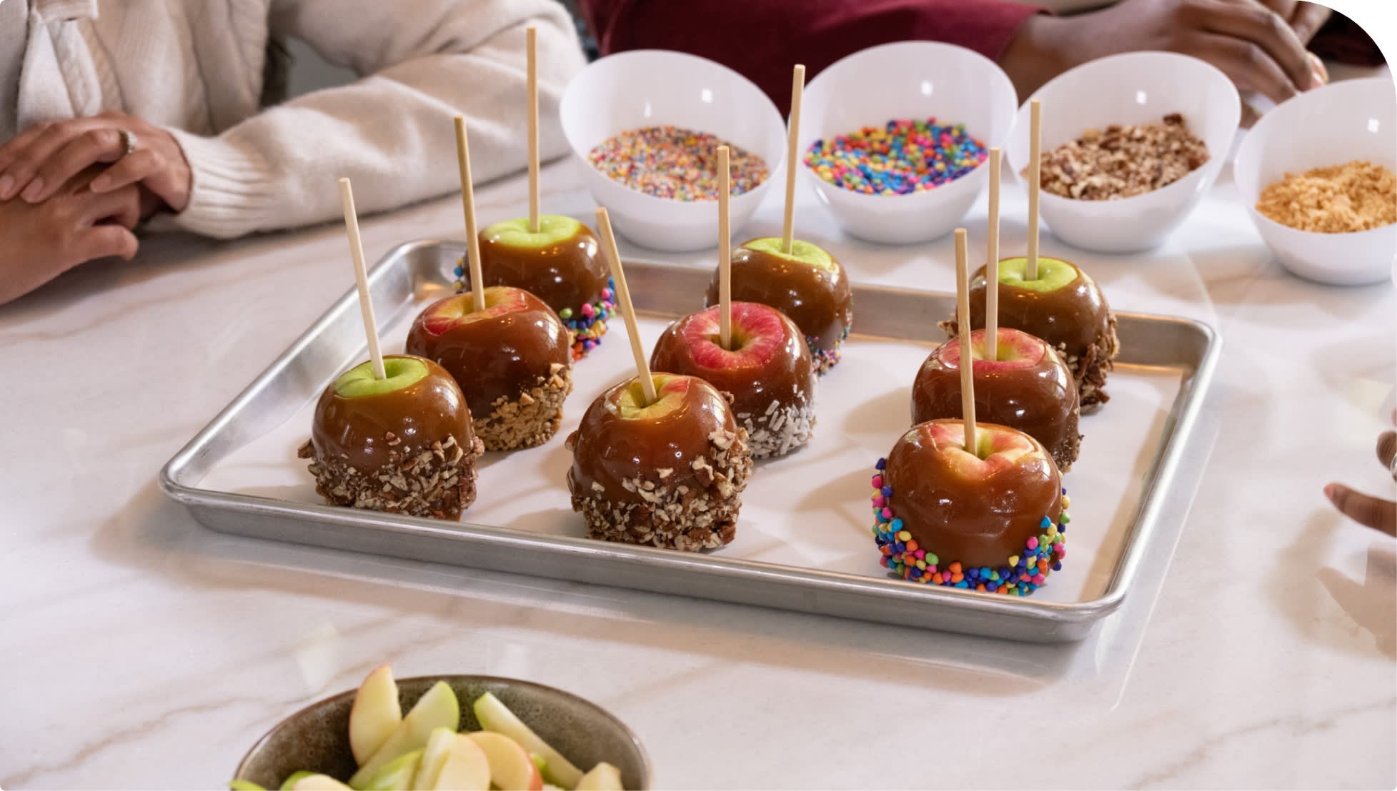 Caramel apples with various toppings on a metal tray on a white marble counter