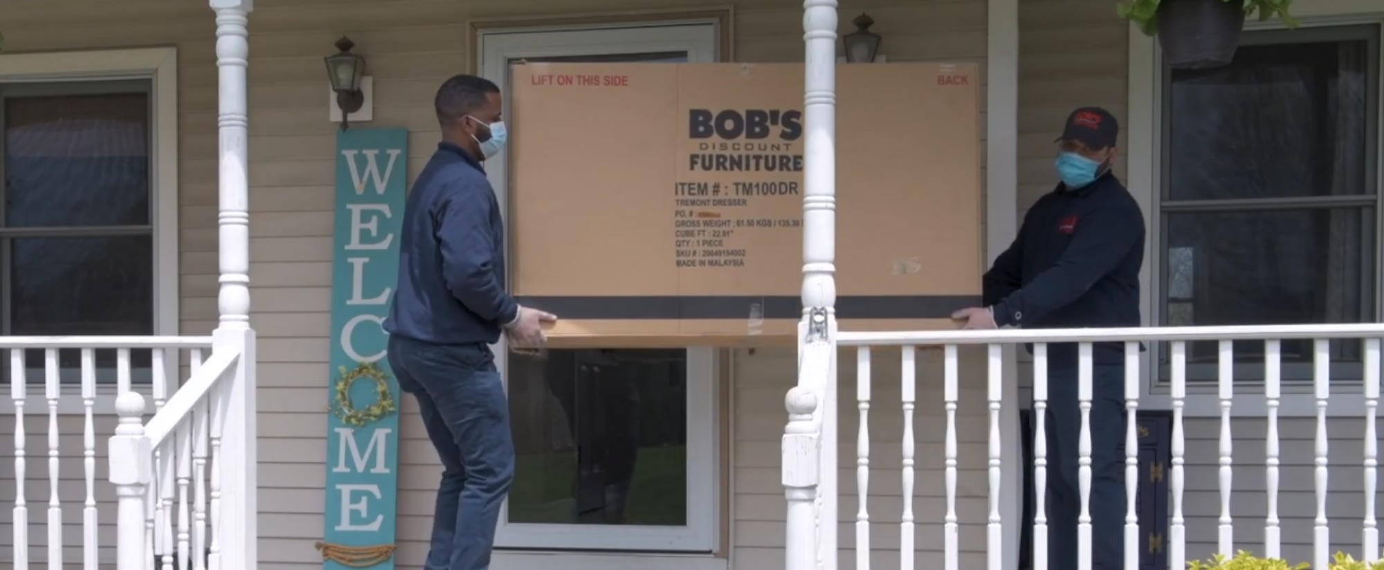 Two delivery workers carry a large Bob's Discount Furniture box onto a porch.