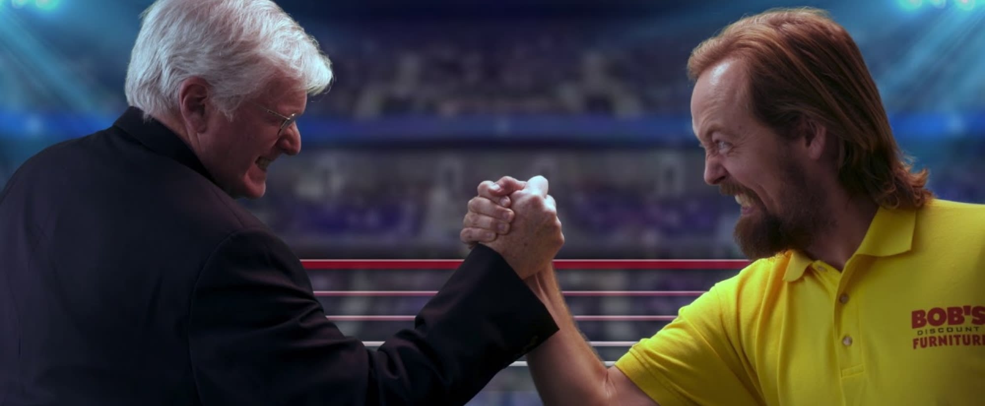 Two men engage in an intense arm-wrestling match in a lively arena. One wears a suit, the other a yellow polo with "Bob's Discount Furniture." Determined expressions.