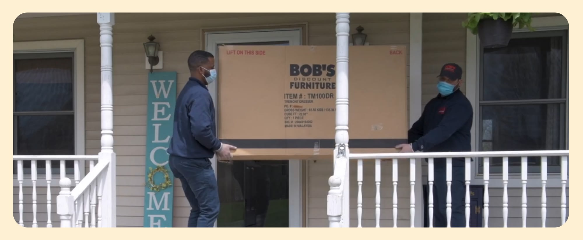 Two delivery workers carry a large Bob's Discount Furniture box onto a porch.
