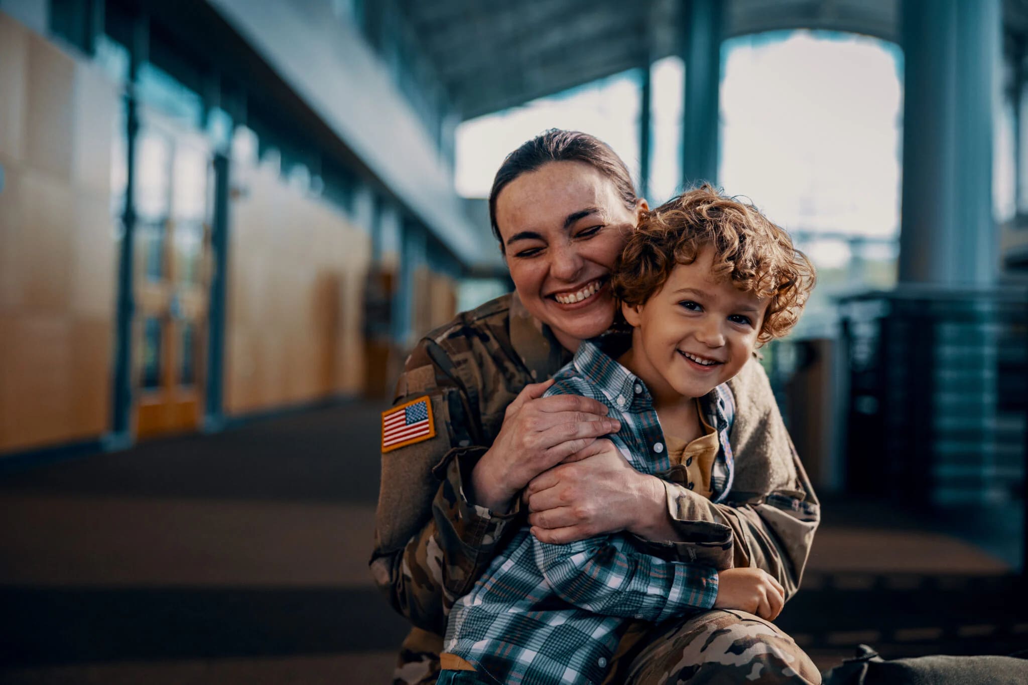 Military personnel in camouflage uniform with an American flag patch embracing a young child wearing a blue plaid shirt inside a spacious, modern building.
