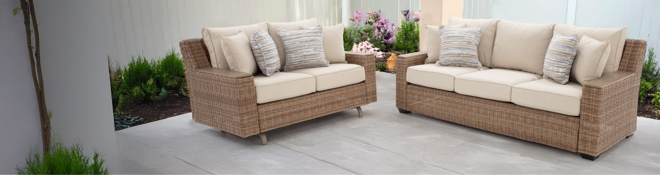 Outdoor patio seating set with beige cushioned wicker loveseat and three-seater sofa, featuring striped accent pillows and wooden armrests on concrete floor.