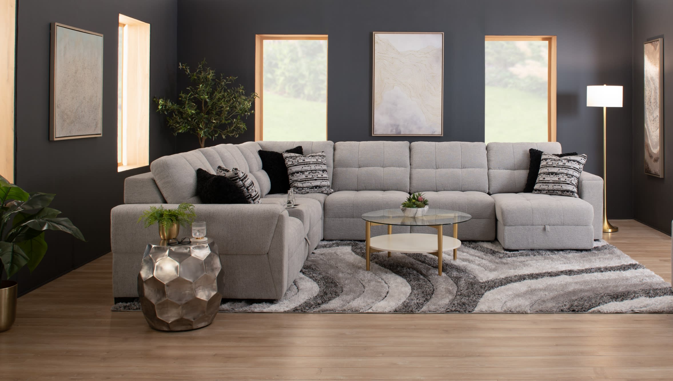 Modern living room with a large light gray fabric sectional sofa, black and patterned throw pillows, a round glass coffee table with gold legs, abstract wall art, and a textured gray and white area rug on light wood flooring.