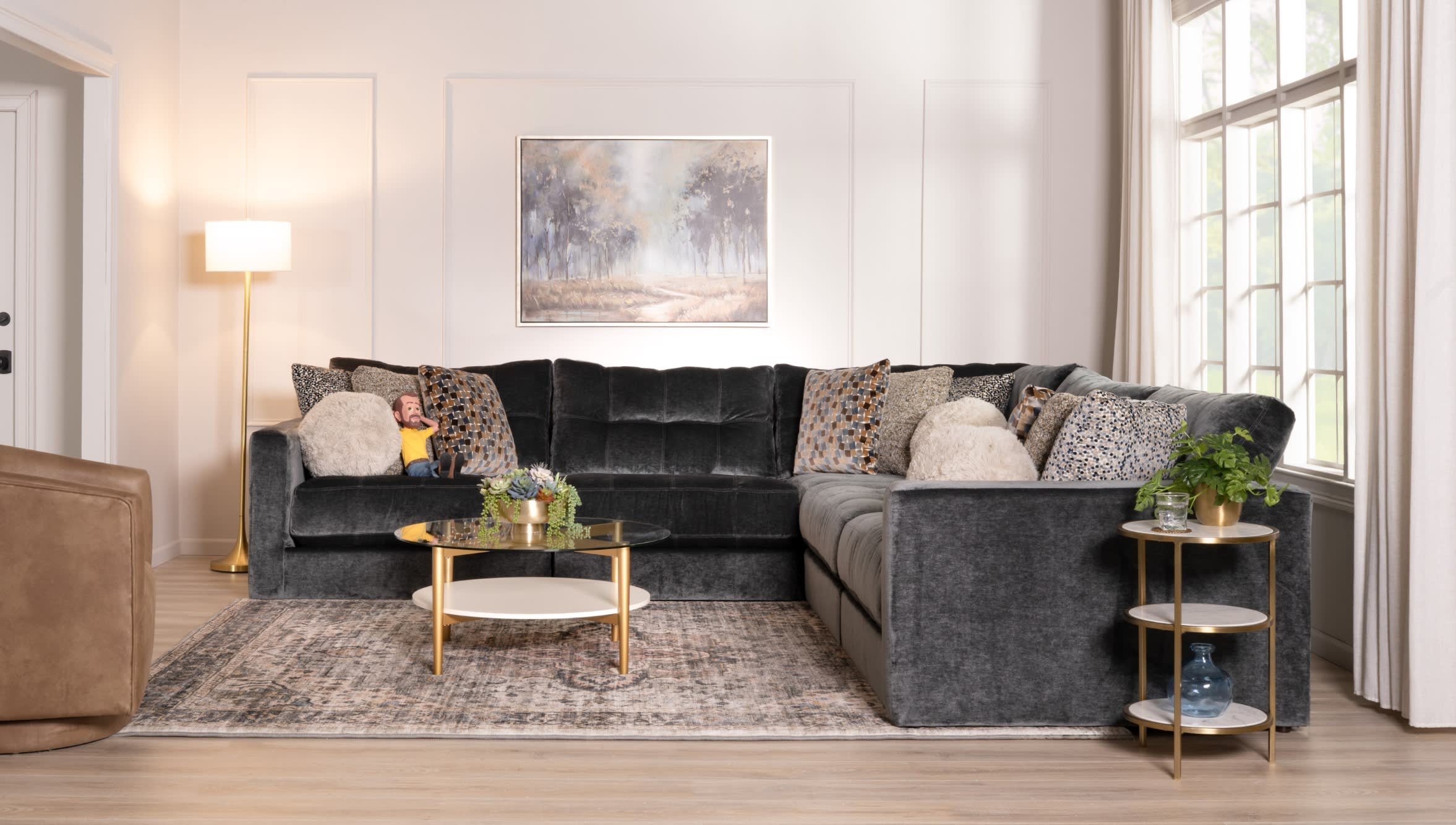 Modern living room with a large dark gray velvet sectional sofa, patterned and textured throw pillows, gold-accented glass coffee table, side table with plant, floor lamp, and large window.