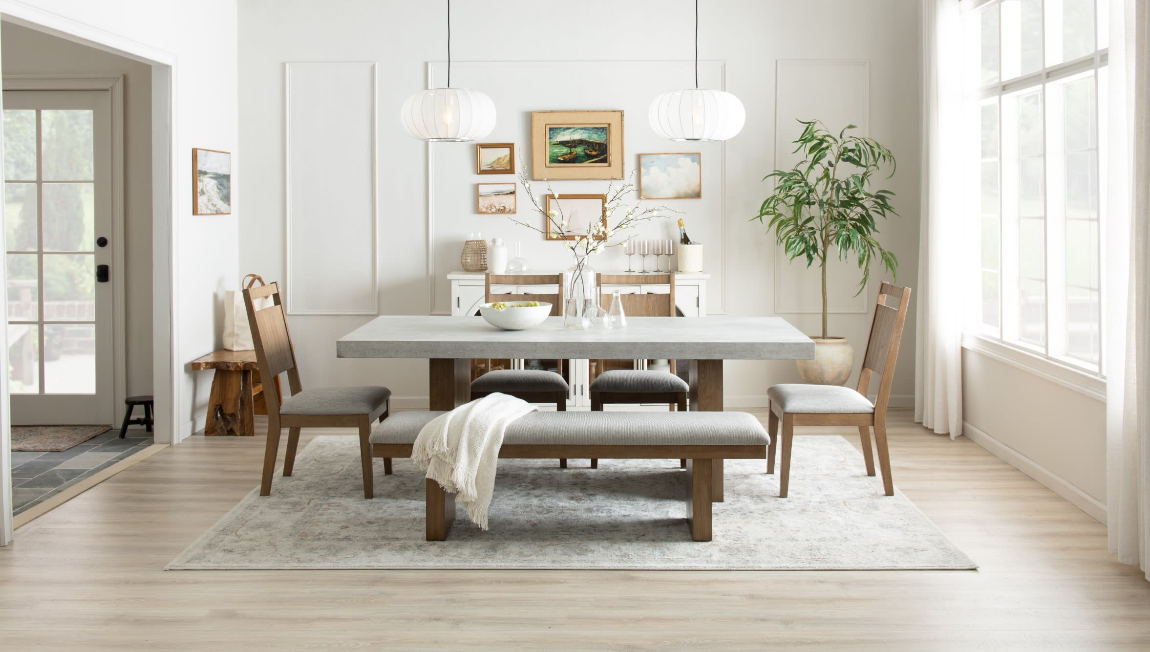 Modern dining room with a rectangular concrete tabletop, wooden chairs and bench with gray cushions, neutral rug, and large window lighting.