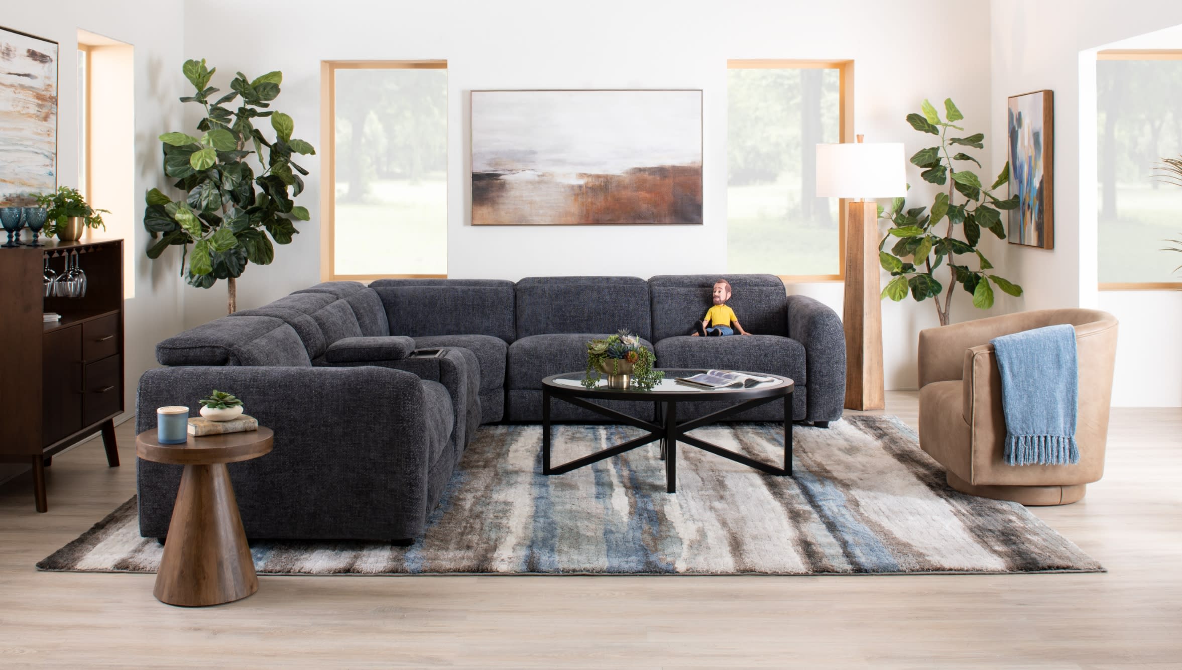 Modern living room with a dark gray fabric sectional sofa, round black metal coffee table, beige swivel chair with blue throw, wooden side table, indoor plants, and abstract wall art.