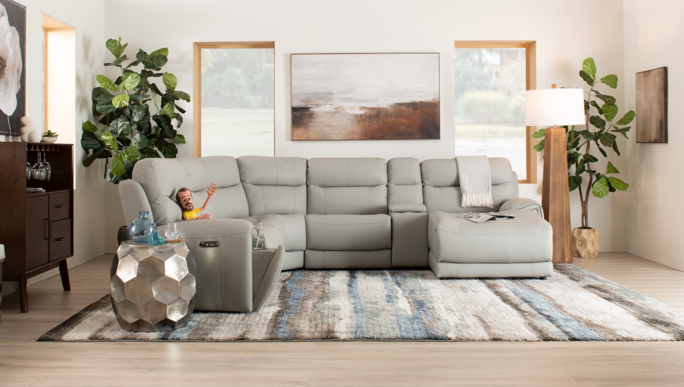 Modern living room with a light gray leather sectional sofa featuring recliners, a textured area rug with blue and beige tones, wooden side table, floor lamp, and green plants.