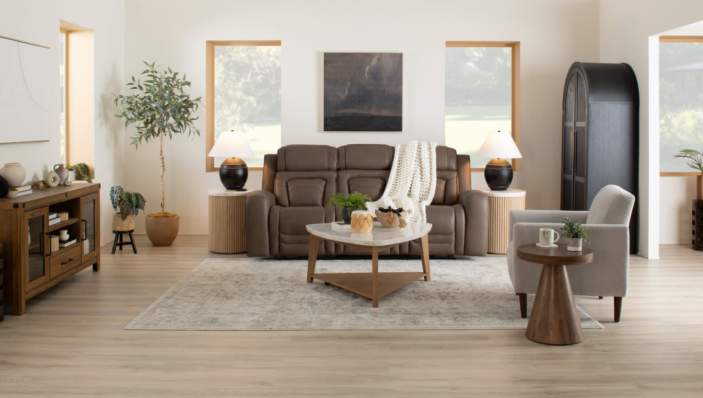 Modern living room with a brown leather recliner sofa, beige armchair, wooden coffee and side tables, decorative plants, and soft area rug.