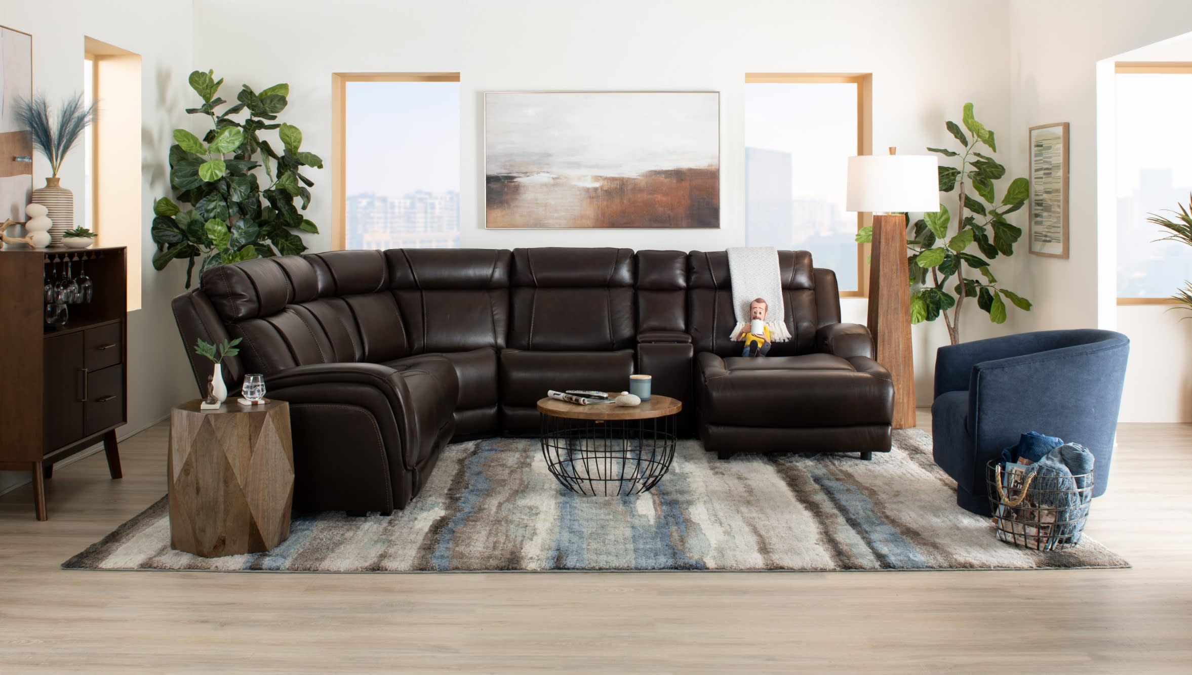 Dark brown leather sectional sofa with reclining seats, wooden geometric side table, round wireframe coffee table, blue upholstered armchair, and floor lamp in a modern living room with large windows and decorative plants.