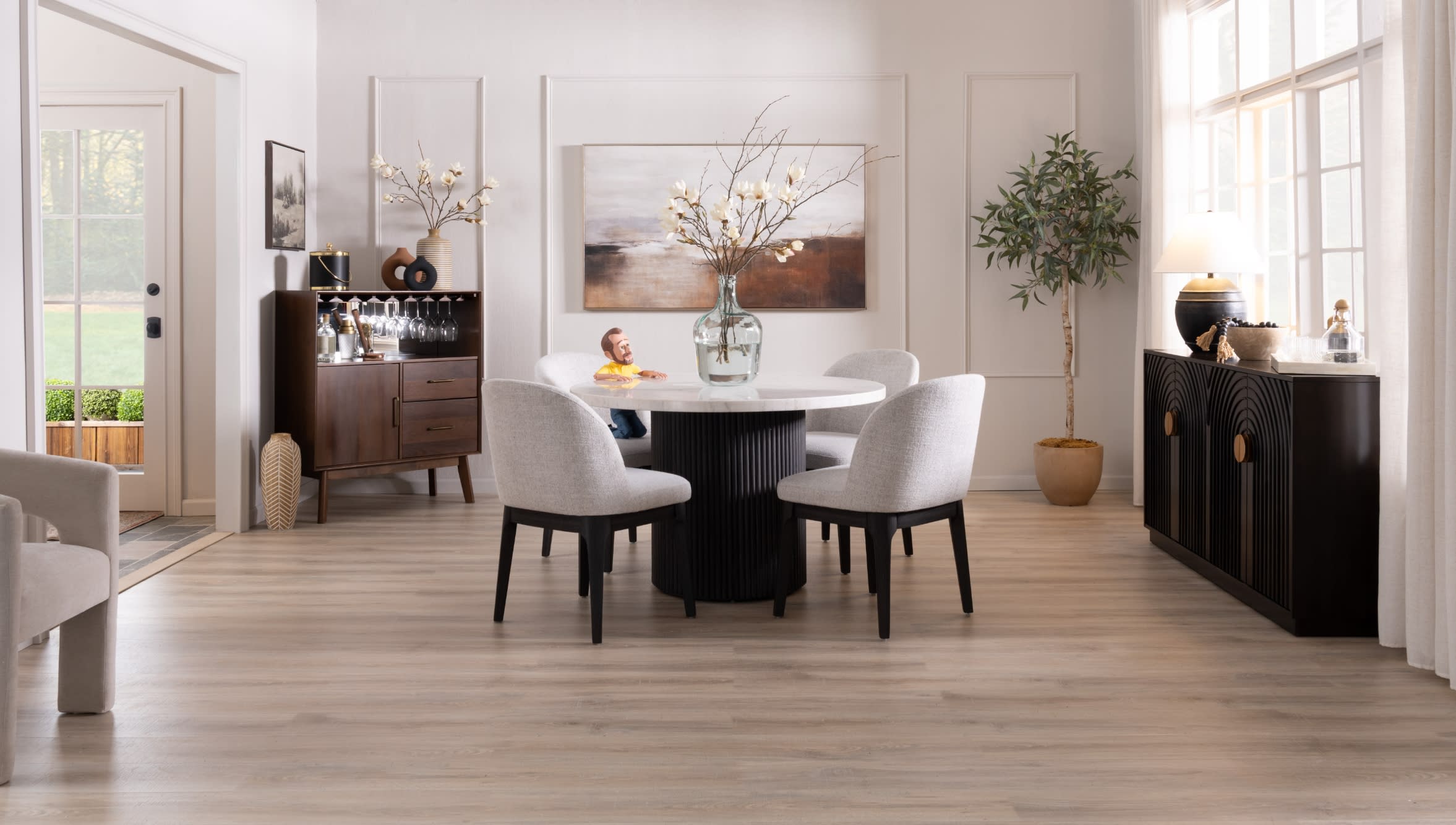 Modern dining room with a round white marble table, four light gray upholstered chairs with black legs, a dark wood sideboard, and large windows.