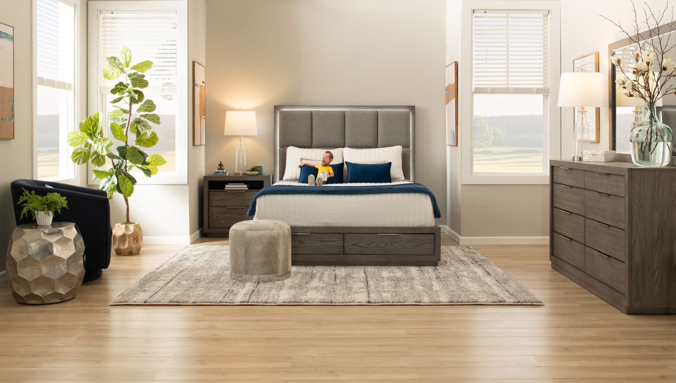 Modern bedroom with a gray upholstered bed, white and navy bedding, wooden nightstands, light wood flooring, and natural light from windows.