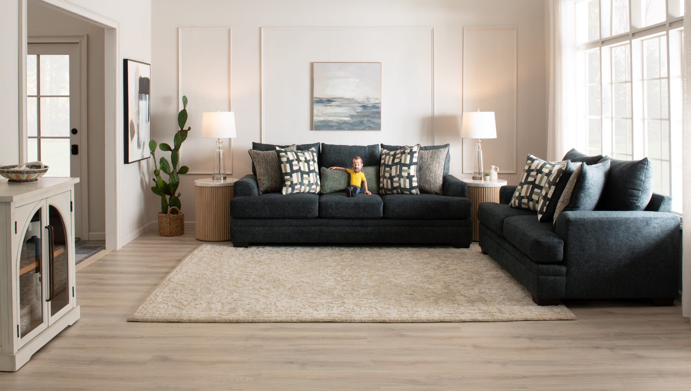 Modern living room with dark gray fabric sofas featuring patterned and textured cushions, light wood flooring, beige area rug, side tables with glass lamps, and large windows providing natural light.