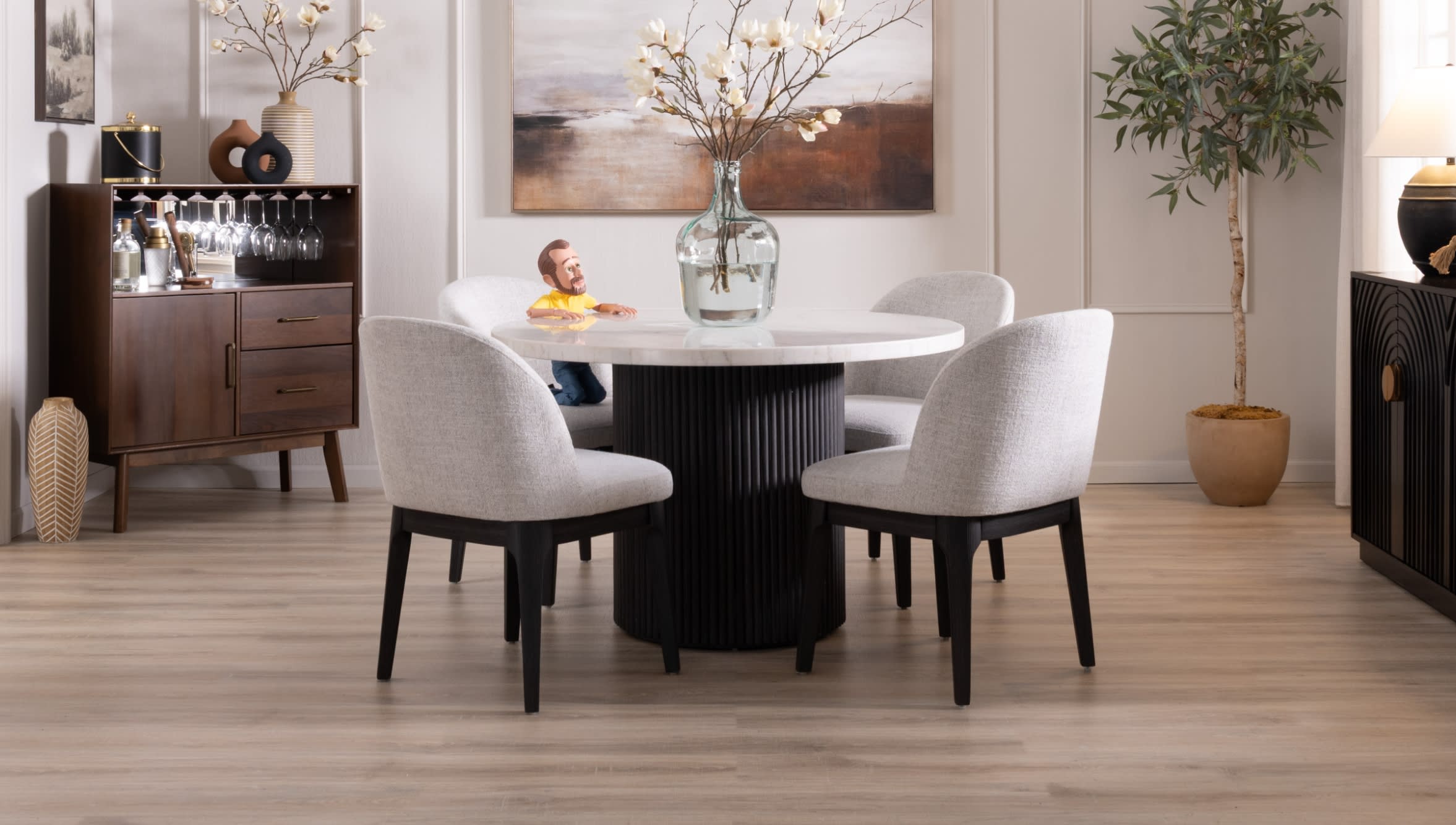 Modern dining room with a round white marble table, four gray upholstered chairs with black legs, a glass vase with flowers, and wooden side cabinets.