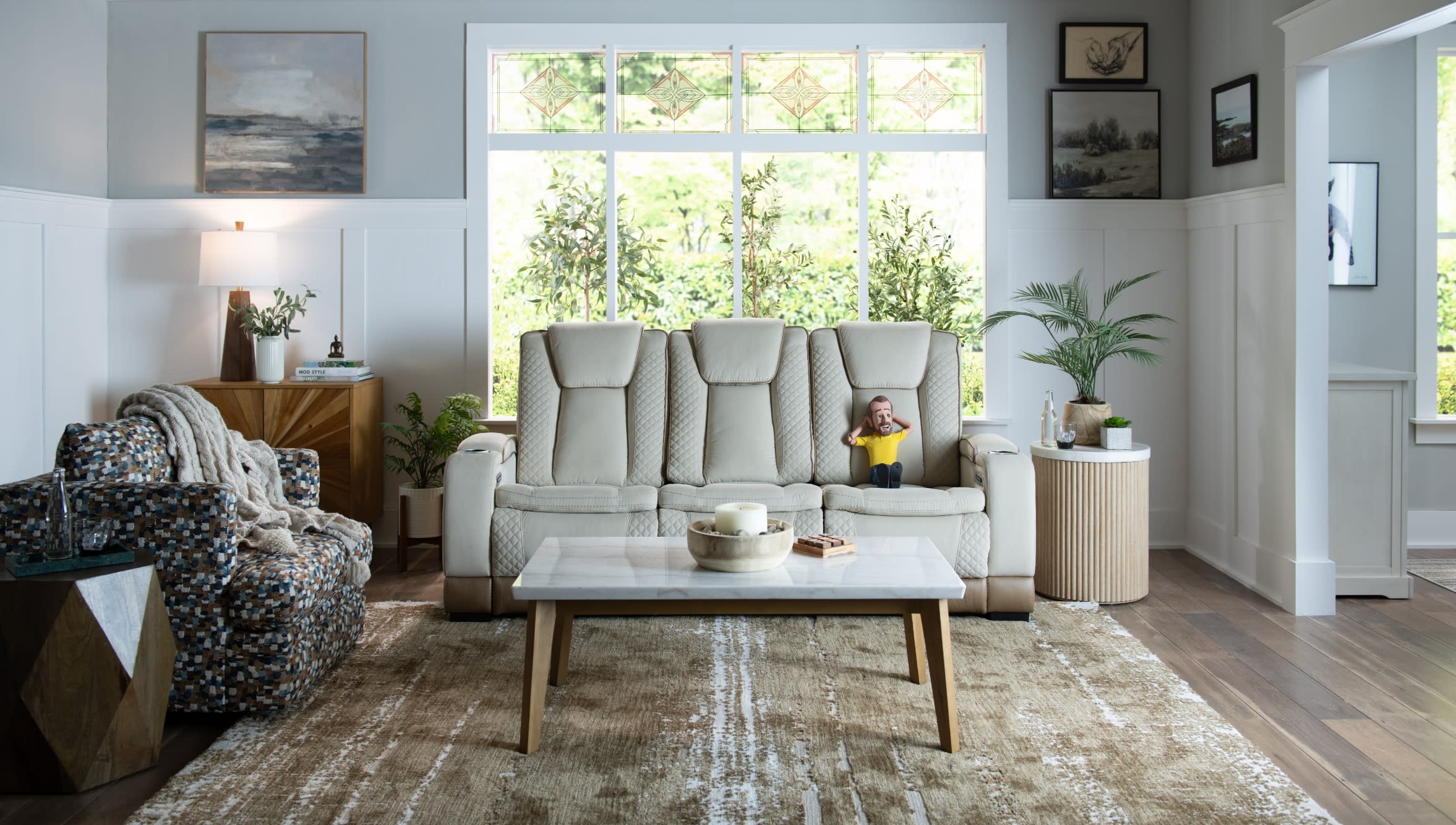 Modern living room with a beige leather recliner sofa, patterned armchair, wooden coffee table with marble top, and large window with greenery outside.