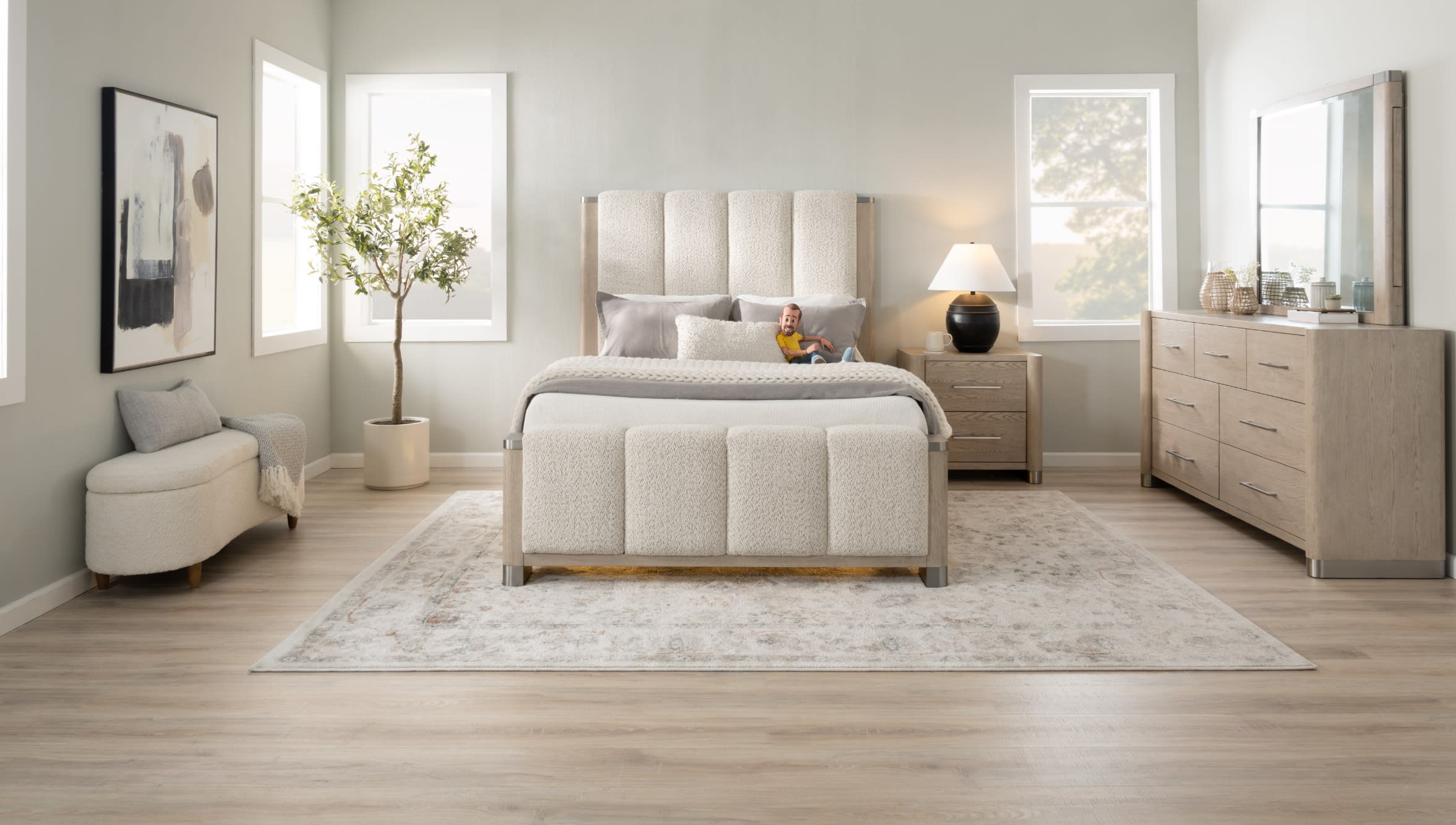 Modern bedroom with beige upholstered bed featuring a textured headboard and footboard, matching nightstand with black lamp, light wood dresser with mirror, abstract wall art, and light wood flooring.
