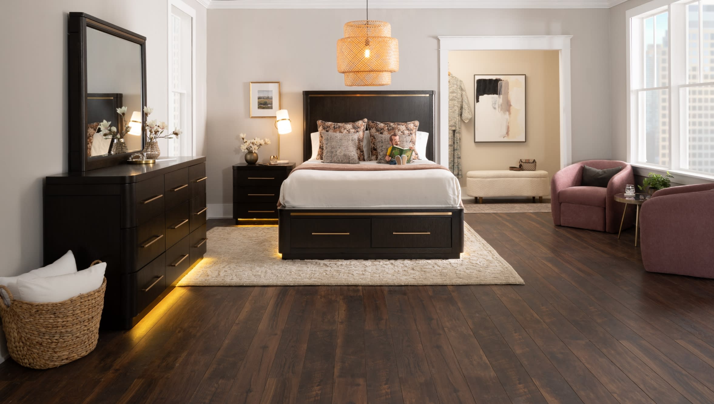 Modern bedroom with dark wood bed frame, matching dresser and nightstand with gold handles, beige rug, and pink armchairs by large windows.