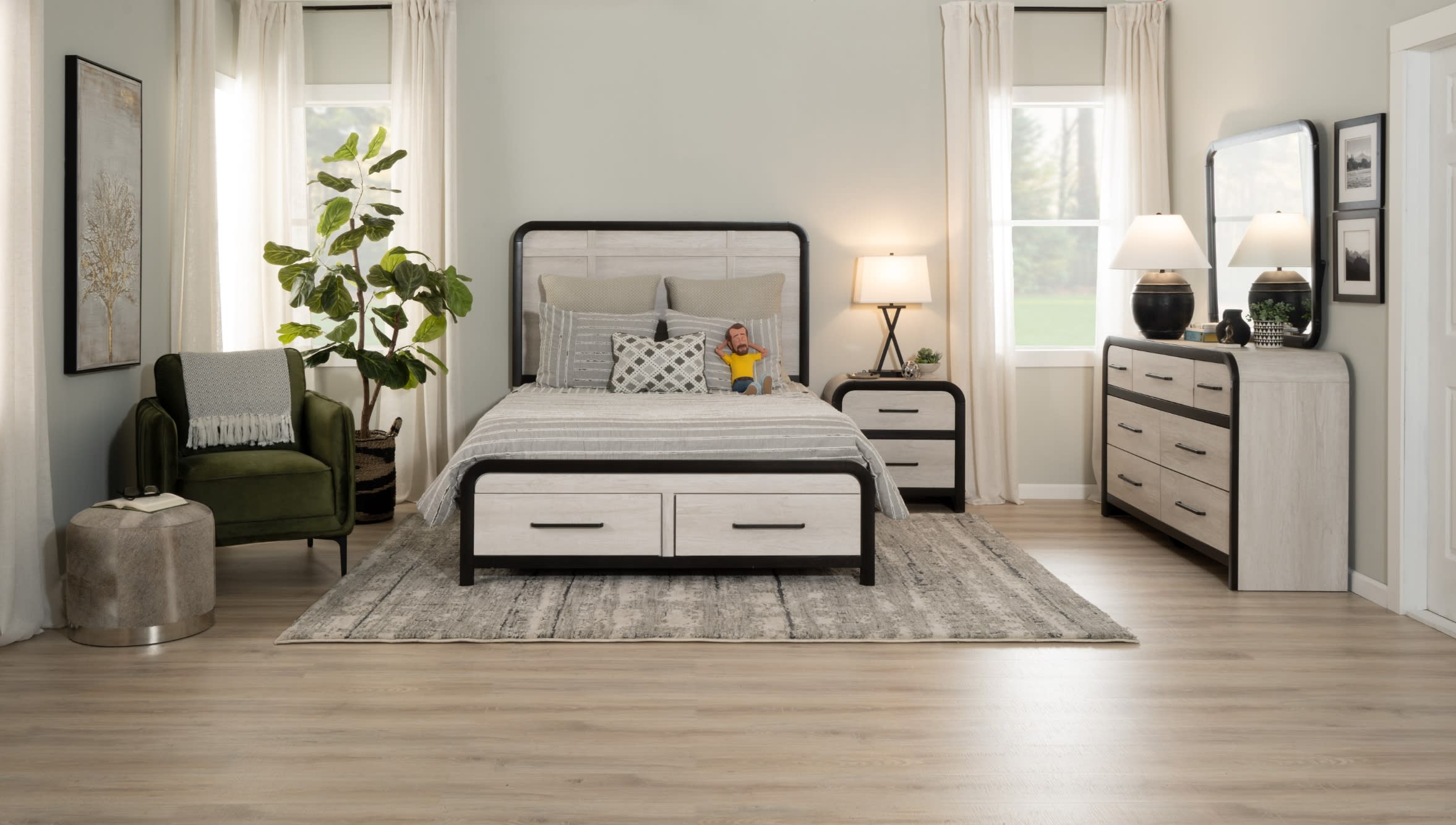 Modern bedroom setup with light wood and black metal bed frame, matching nightstand and dresser, gray patterned rug, green armchair, and neutral decor.