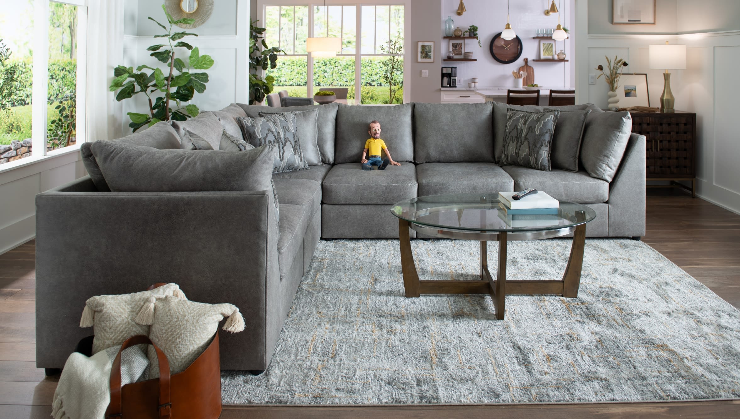 Modern living room with a large gray fabric sectional sofa featuring patterned throw pillows, a glass-top wooden coffee table, and a textured area rug.