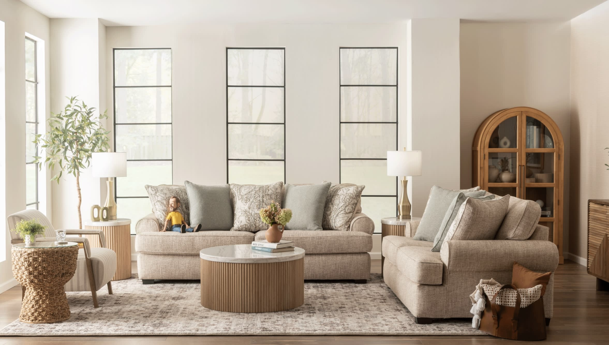 Modern living room with beige fabric sofas, patterned and solid cushions, round wooden coffee table with marble top, large windows, and natural light.