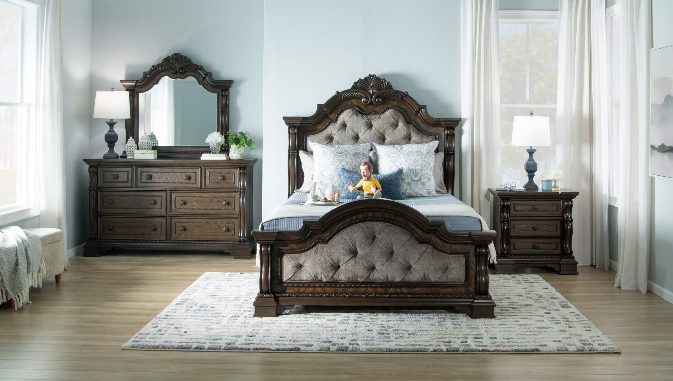 Traditional dark wood bedroom set with tufted upholstered headboard and footboard, matching dresser with mirror, and nightstand, styled in a bright room with light wood flooring and neutral decor.