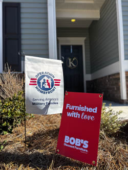 Two yard flags displayed in front of a house: a white flag with the Operation Homefront logo and text "Serving America's Military Families," and a red flag with white text "Furnished with love BOB'S Discount Furniture mybobs.com," promoting military family support and furniture discount.